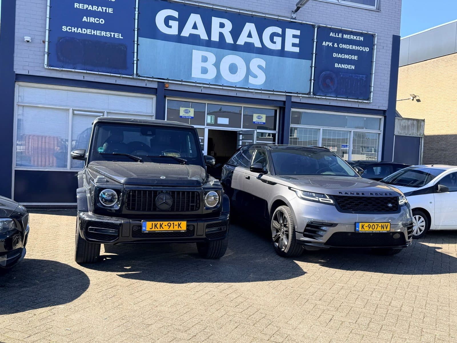 De voorgevel van Garage Bos in de zon, met de gevelreclame voor APK, onderhoud, diagnose en banden en twee auto's op het voorterrein